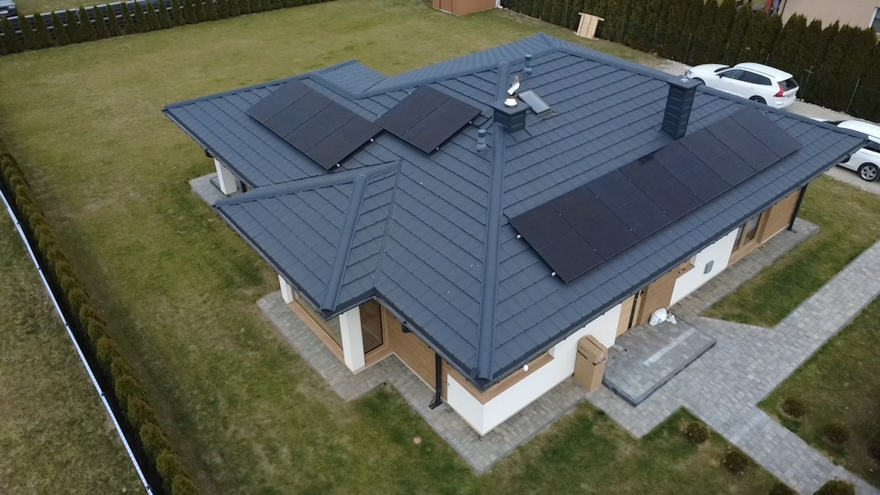 Dom jednorodzinny z ciemną dachówką i panelami słonecznymi na dachu, widok z góry. Na trawniku zaparkowane dwa samochody osobowe.