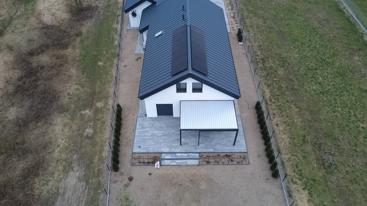 Widok z drona na dom z ciemną dachówką i panelami słonecznymi. Jasna elewacja, szara kostka brukowa na tarasie i zadbany ogród z młodymi tujami wzdłuż ogrodzenia.