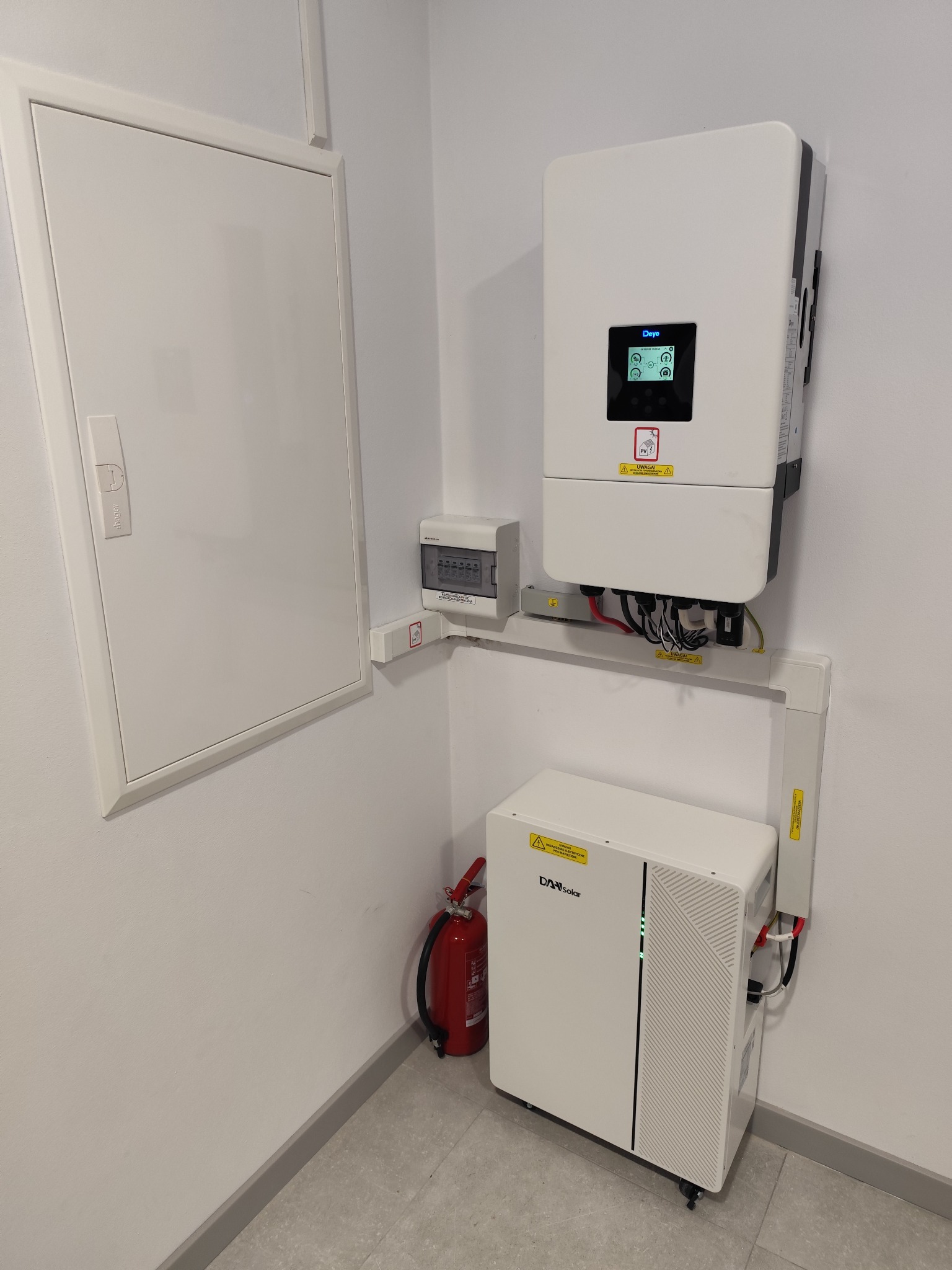 Biała instalacja z inwerterem i akumulatorem Daye Solar na ścianie, obok skrzynka elektryczna i gaśnica. Widoczne kable i oznaczenia ostrzegawcze.