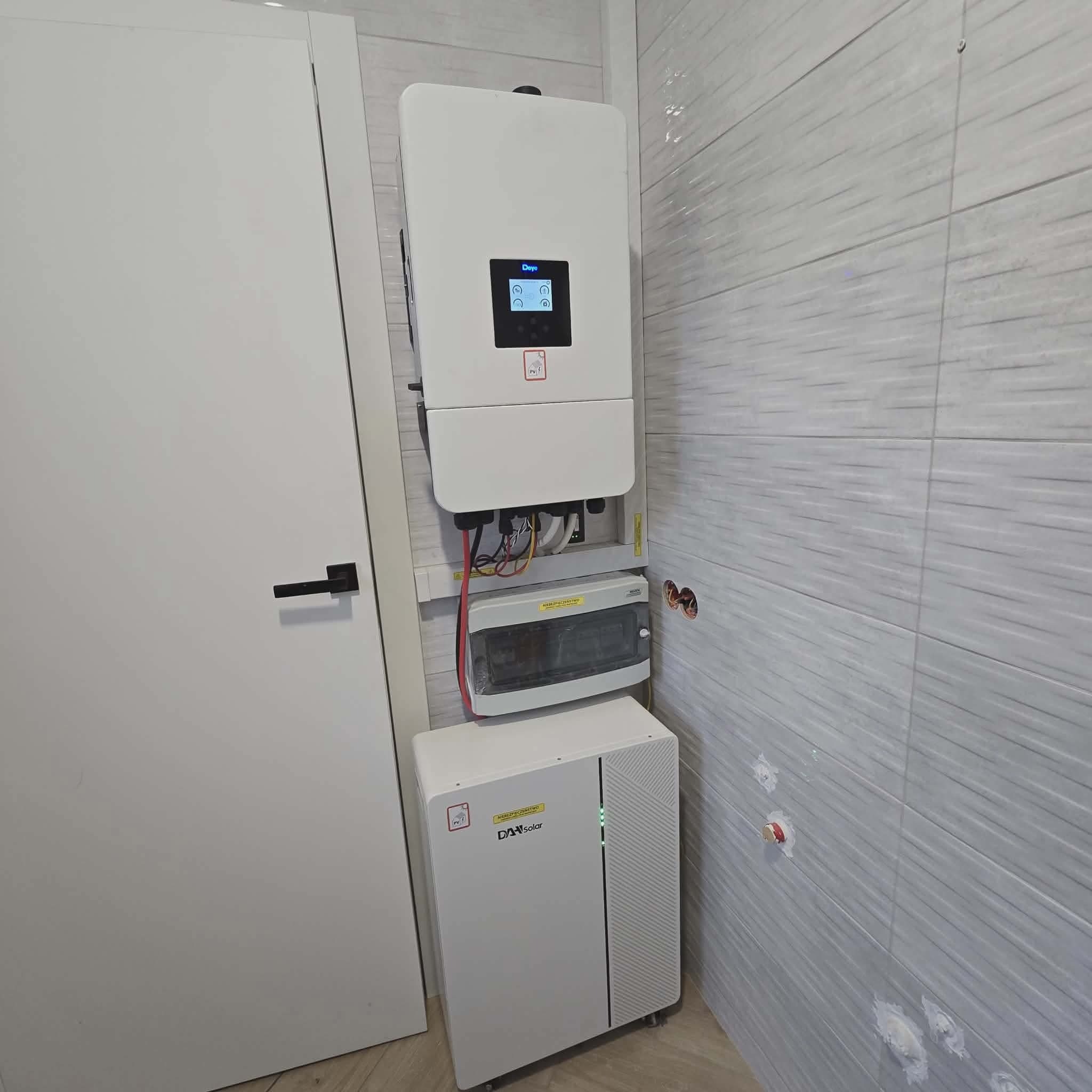 Biały inwerter fotowoltaiczny Deye i magazyn energii DAH Solar zamontowane na ścianie z szarymi płytkami wewnątrz pomieszczenia, obok białe drzwi.