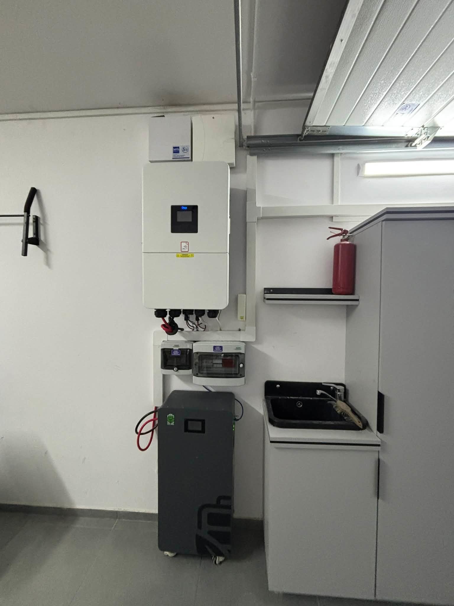 Zamontowany, kompletny system zasilania awaryjnego w garażu: biały inwerter, szary akumulator, skrzynka bezpiecznikowa, okablowanie. Gaśnica na półce.