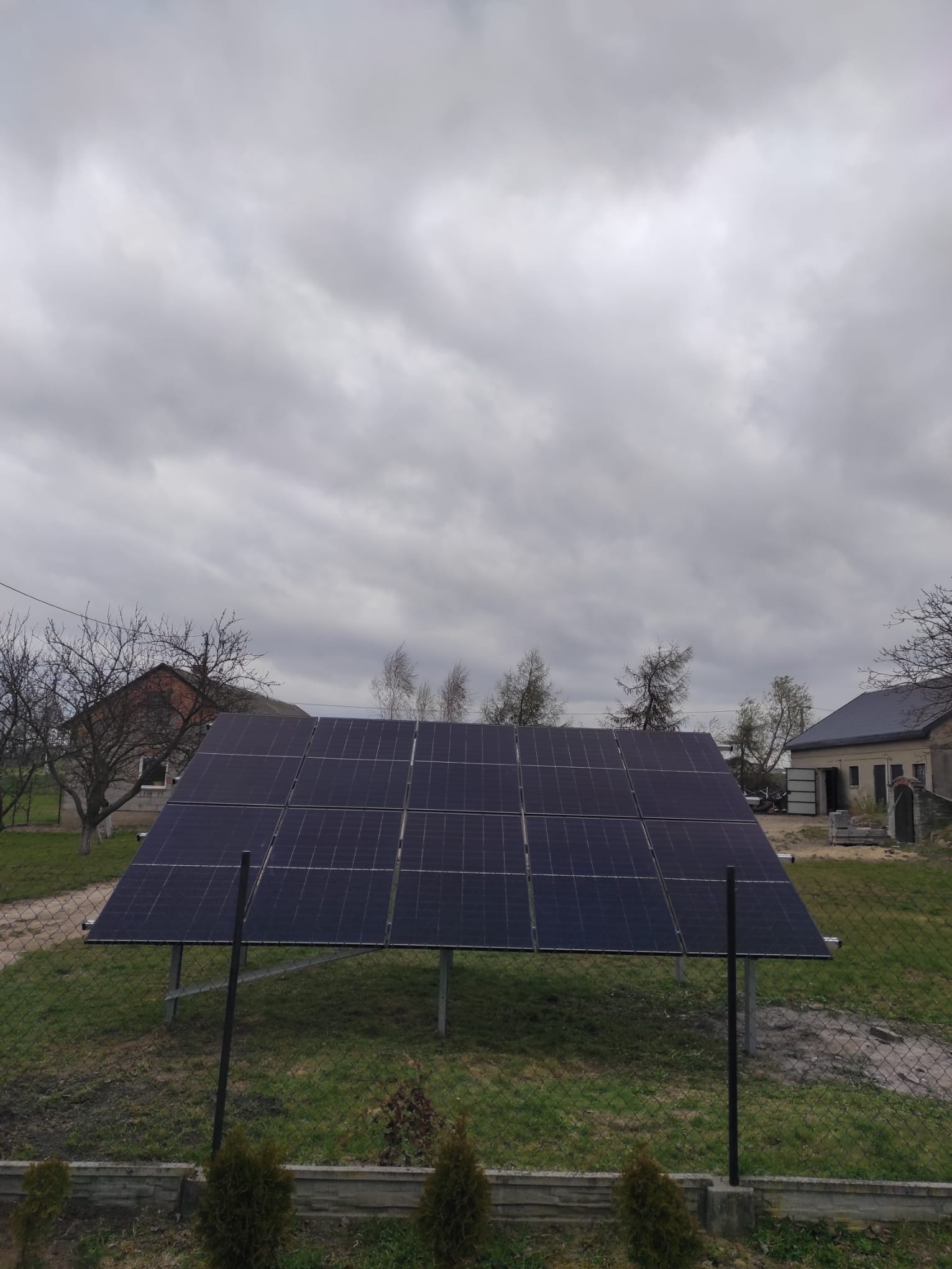 Panel słoneczny na metalowej konstrukcji, widoczny zza ogrodzenia. W tle budynki i pochmurne niebo. Instalacja w wiejskim otoczeniu.