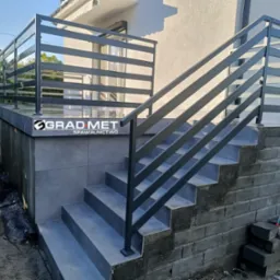 Zewnętrzne schody z szarą metalową balustradą poziomą, prowadzące do wejścia domu, z widocznym logo firmy na balustradzie i materiałami budowlanymi obok schod&oacute;w.