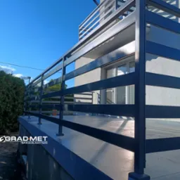 Nowoczesna, pozioma balustrada metalowa w kolorze grafitowym, zamontowana na balkonie domu z widokiem na błękitne niebo i zieleń drzew.