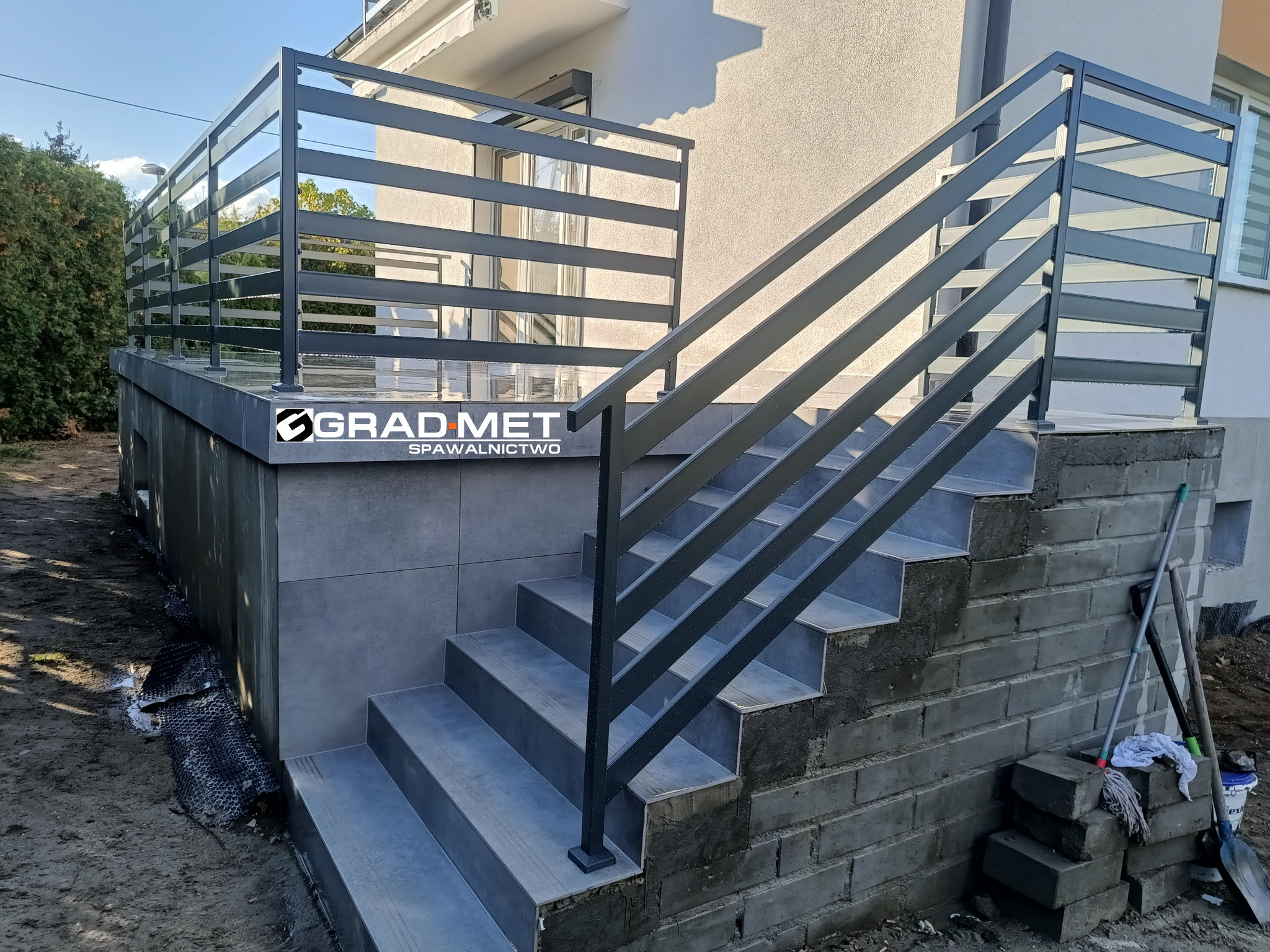 Zewnętrzne schody z szarą metalową balustradą poziomą, prowadzące do wejścia domu, z widocznym logo firmy na balustradzie i materiałami budowlanymi obok schodów.