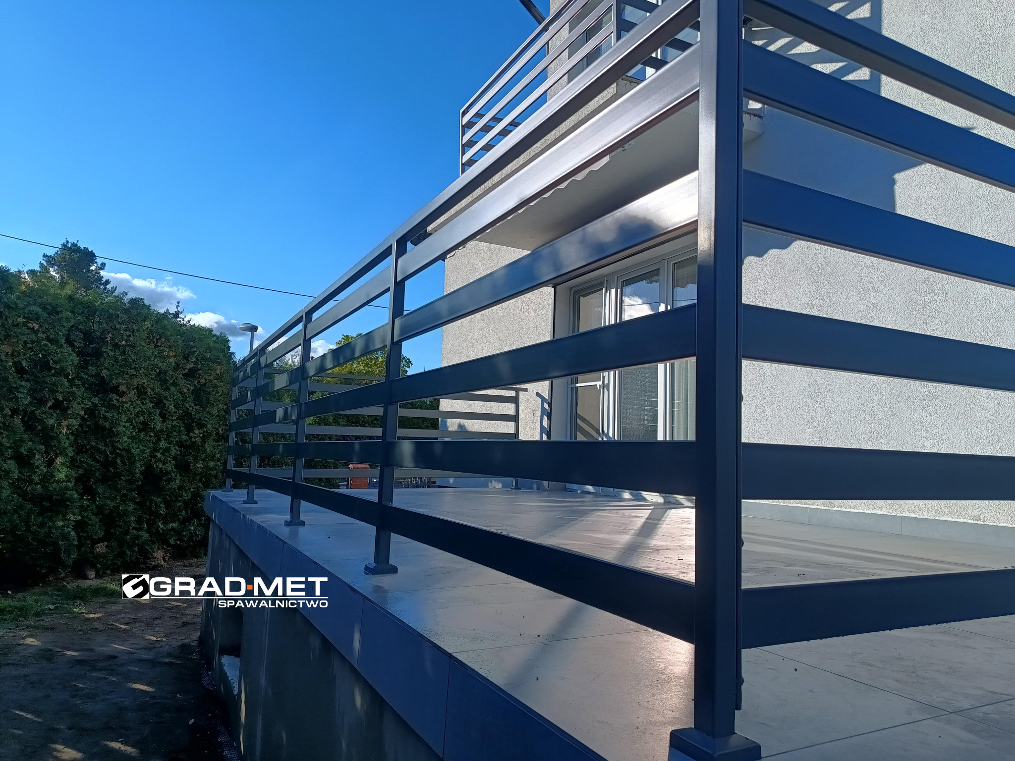 Nowoczesna, pozioma balustrada metalowa w kolorze grafitowym, zamontowana na balkonie domu z widokiem na błękitne niebo i zieleń drzew.