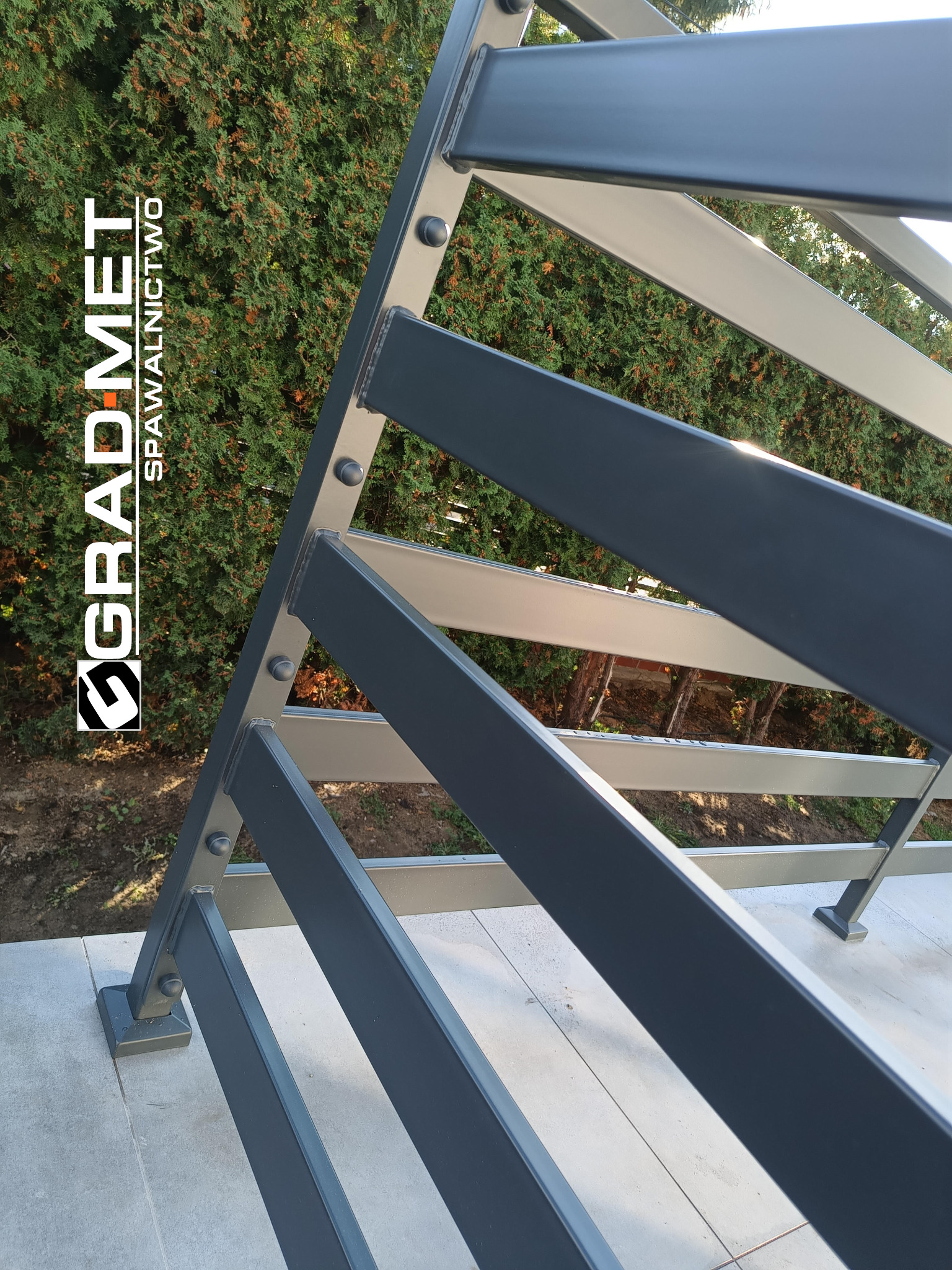 Metalowa konstrukcja w kolorze grafitowym z widocznym logo firmy GRADMET, na tle zielonych krzewów.