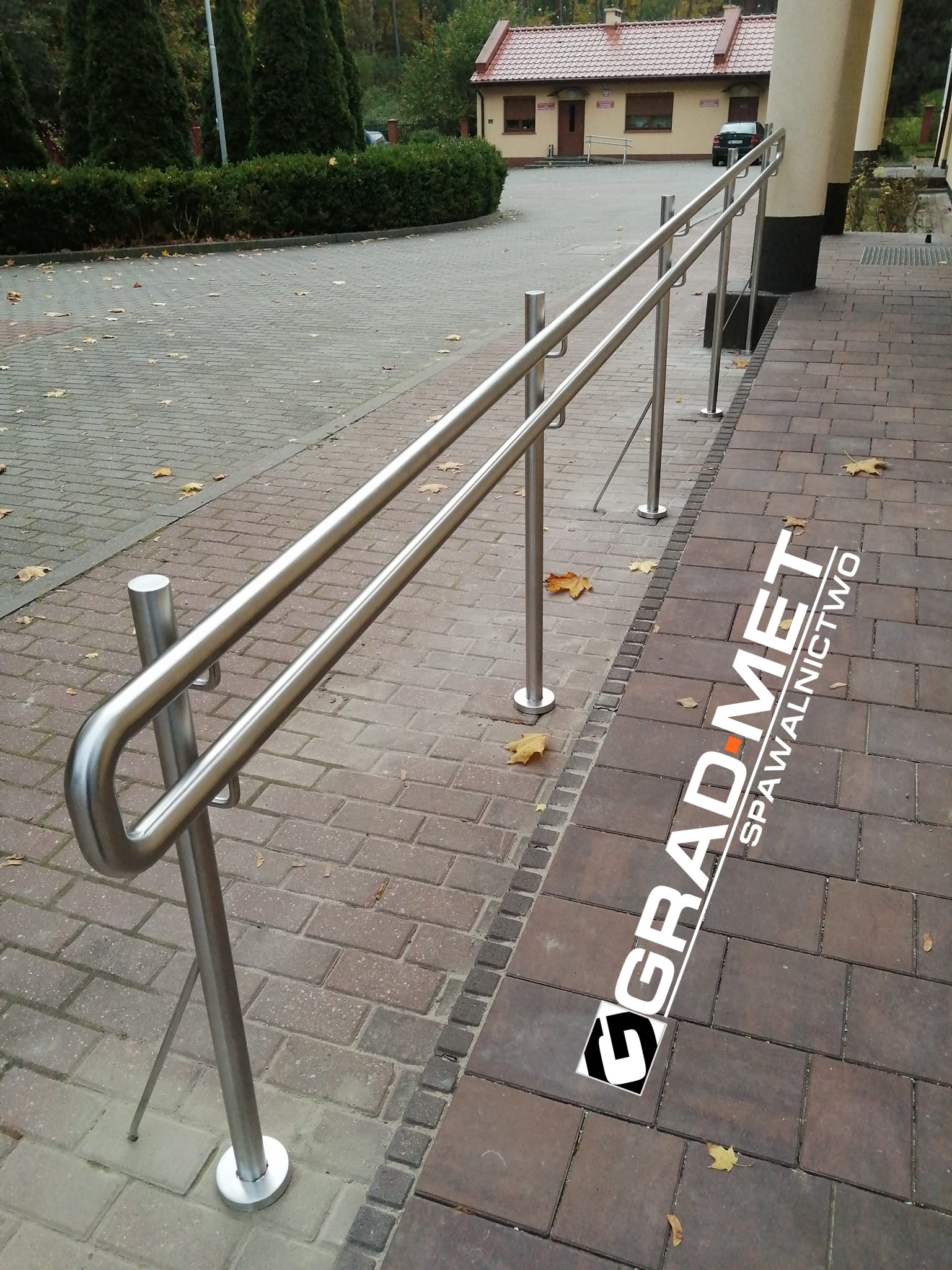 Zewnętrzna poręcz ze stali nierdzewnej zamontowana przy schodach prowadzących do budynku, widoczny bruk i logo firmy na kostce.