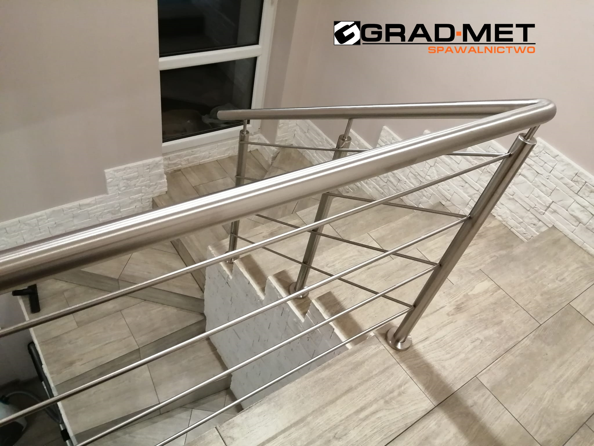 Nowoczesna balustrada ze stali nierdzewnej z poziomymi prętami na schodach wyłożonych płytkami imitującymi drewno, widok z góry, z logo firmy w prawym górnym rogu.