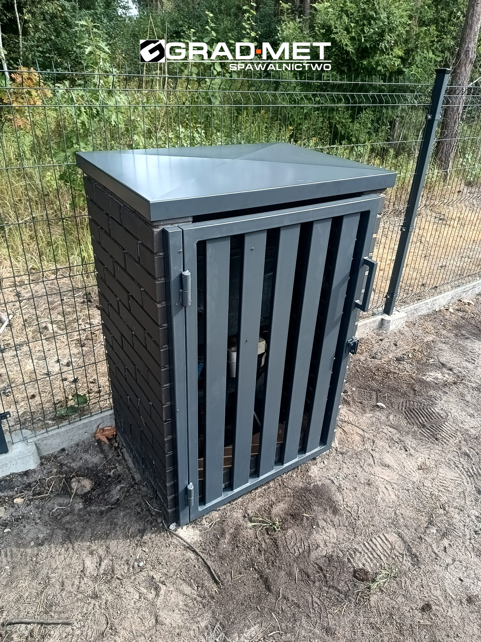 Szara, metalowa obudowa na licznik z ceglaną podstawą i metalowymi drzwiczkami na tle siatki ogrodzeniowej i zieleni.