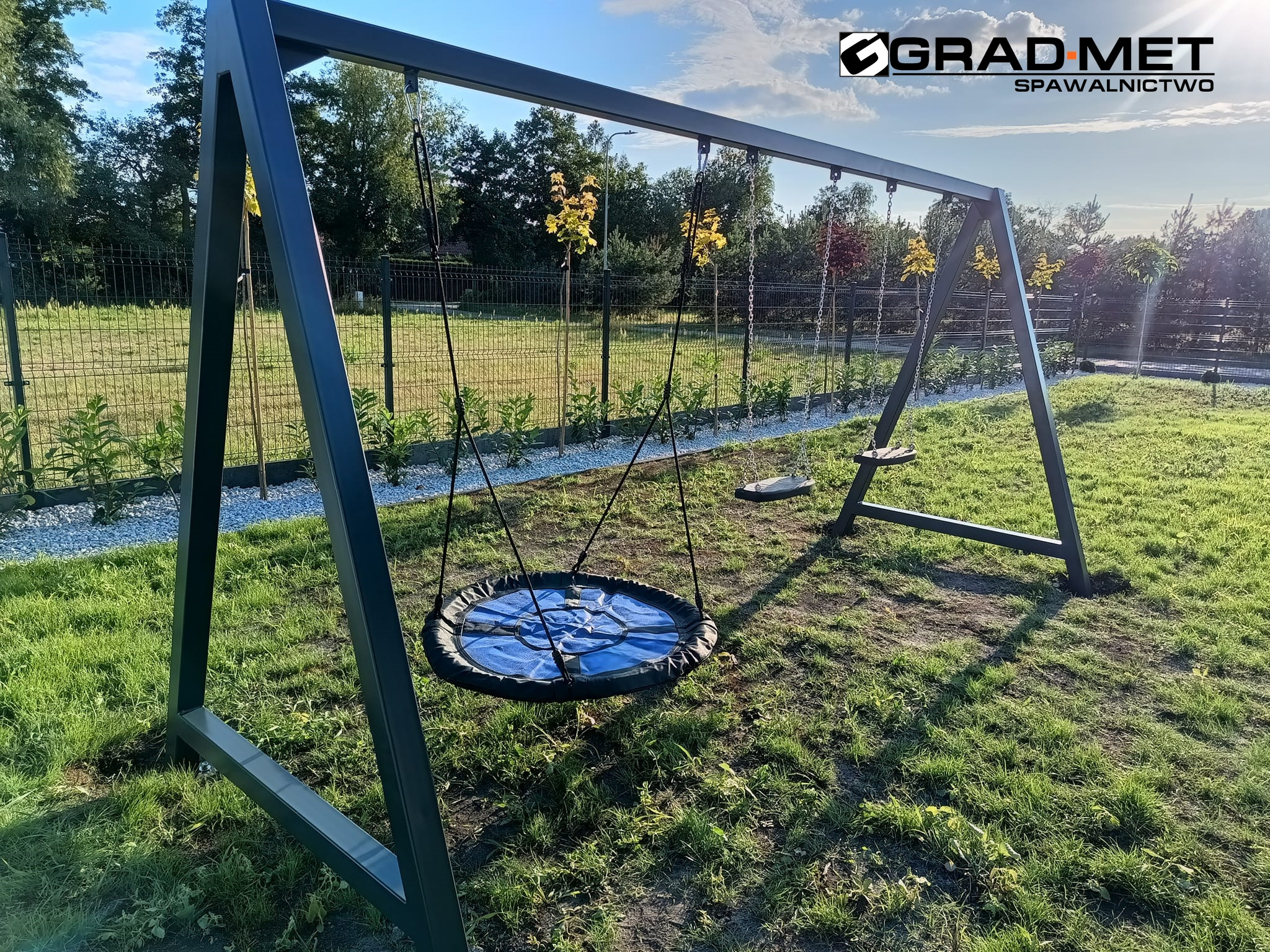 Metalowa huśtawka z okrągłym siedziskiem i standardowym siedziskiem, widoczna na tle ogrodzenia i drzew, z logo firmy Grad-Met Spawalnictwo w prawym górnym rogu.