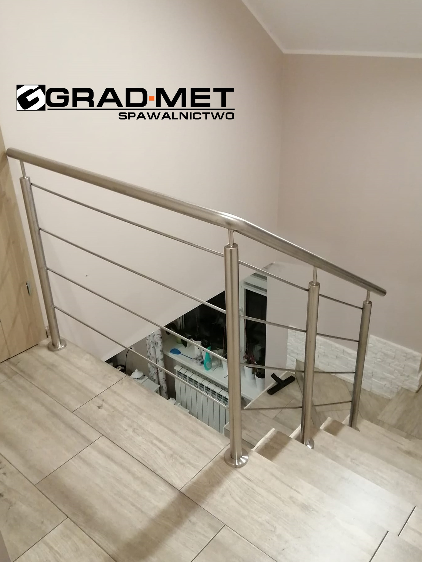 Widok z góry na schody wykończone płytkami imitującymi drewno, zabezpieczone nowoczesną, minimalistyczną poręczą ze stali nierdzewnej z poziomymi prętami, w tle widoczne okno i grzejnik, na ścianie...