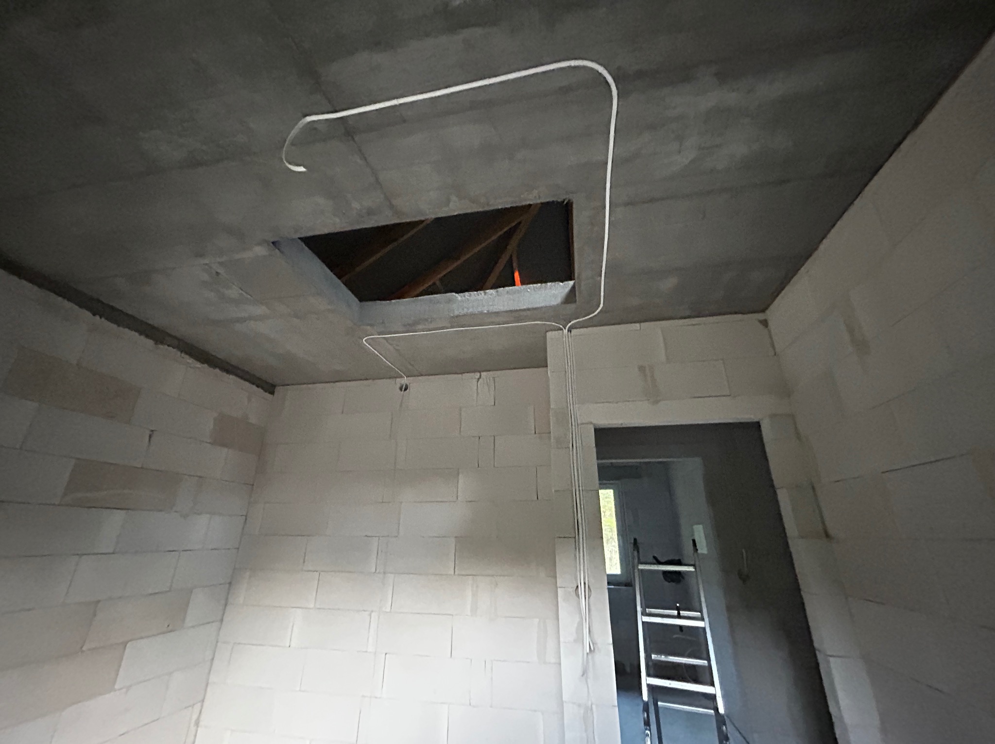 Surowy stan wnętrza z widocznymi przewodami elektrycznymi prowadzonymi po suficie i ścianie z pustaków, otwór w stropie z widoczną konstrukcją dachu, drabina w tle.