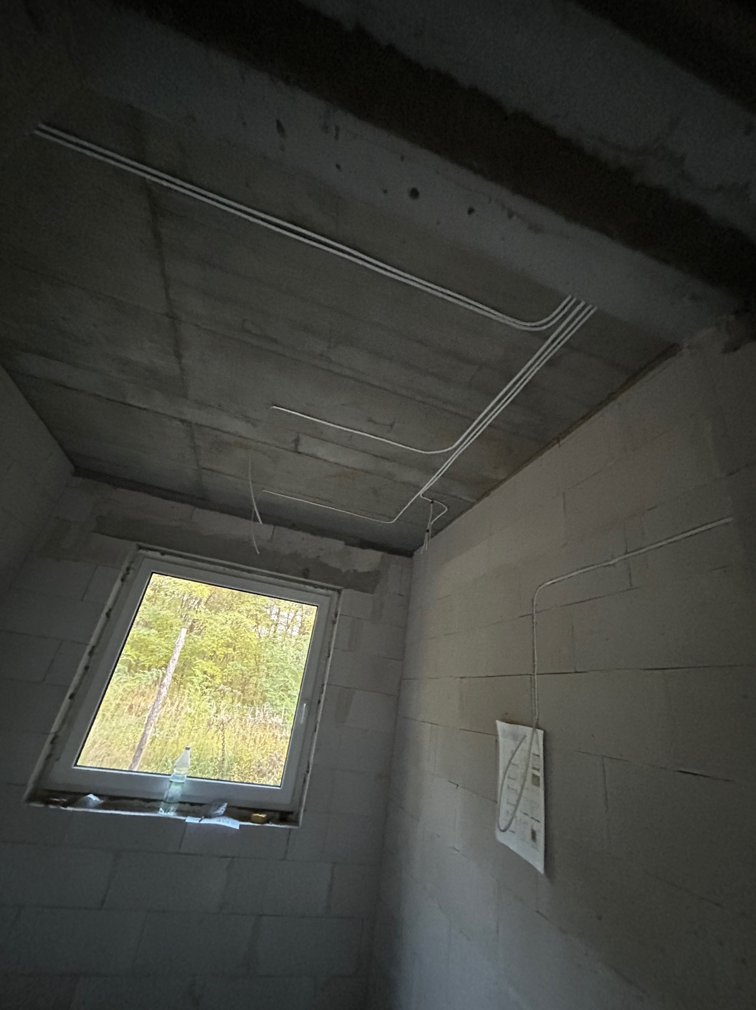 Wnętrze surowego pomieszczenia z widocznymi przewodami elektrycznymi poprowadzonymi po suficie i ścianie, okno z widokiem na zieleń, butelka wody na parapecie, niedokończone ściany z pustaków.
