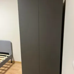 Zamknięta, dwudrzwiowa szafa w odcieniach szarości, z widocznym fragmentem szarego łóżka z drewnianym stelażem, ustawiona na jasnej podłodze z paneli.