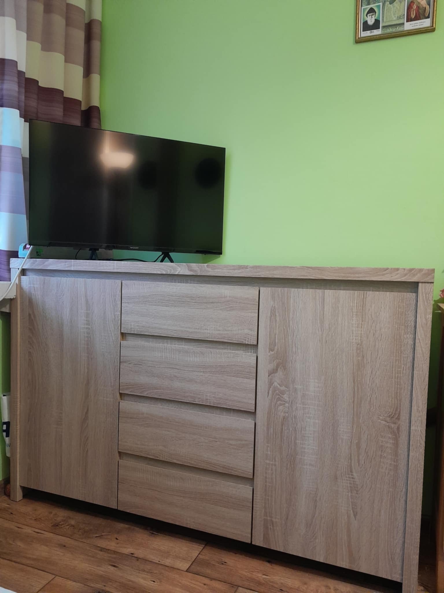 Jasna komoda z trzema szufladami i szafką, ustawiona na drewnianej podłodze, z telewizorem na blacie i zieloną ścianą w tle.