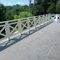 Biała balustrada z krzyżowym wzorem na tarasie z widokiem na zieleń, wyraźne cienie w słoneczny dzień.