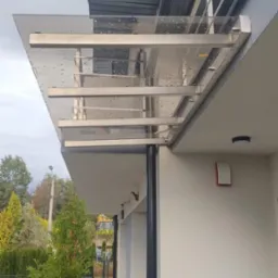 Zadaszenie nad wejściem do domu, konstrukcja ze stali nierdzewnej i przezroczystego tworzywa, widoczne krople deszczu na powierzchni, w tle drzewa i fragment ogrodzenia panelowego.