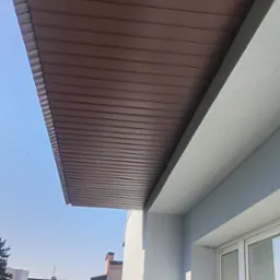 Podbitka dachowa z brązowych paneli PCV zamontowana na balkonie budynku mieszkalnego, widok z dołu na tle błękitnego nieba i zabudowy miejskiej.