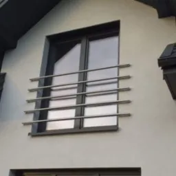 Zewnętrzna balustrada okienna z polerowanej stali nierdzewnej, składająca się z czterech poziomych prętów zamocowanych na elewacji budynku z ciemną stolarką okienną i grafitowym dachem.