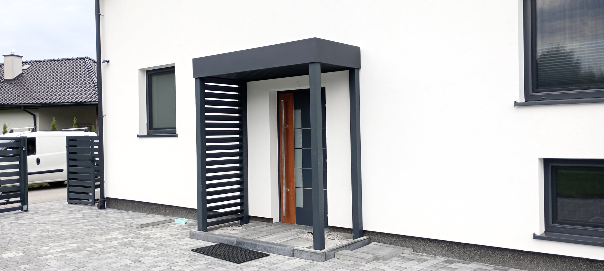 Nowoczesne zadaszenie nad wejściem do domu, wykonane z grafitowych profili metalowych, z elementami ażurowymi po lewej stronie, dopasowane kolorystycznie do stolarki okiennej i bramy wjazdowej w tle.