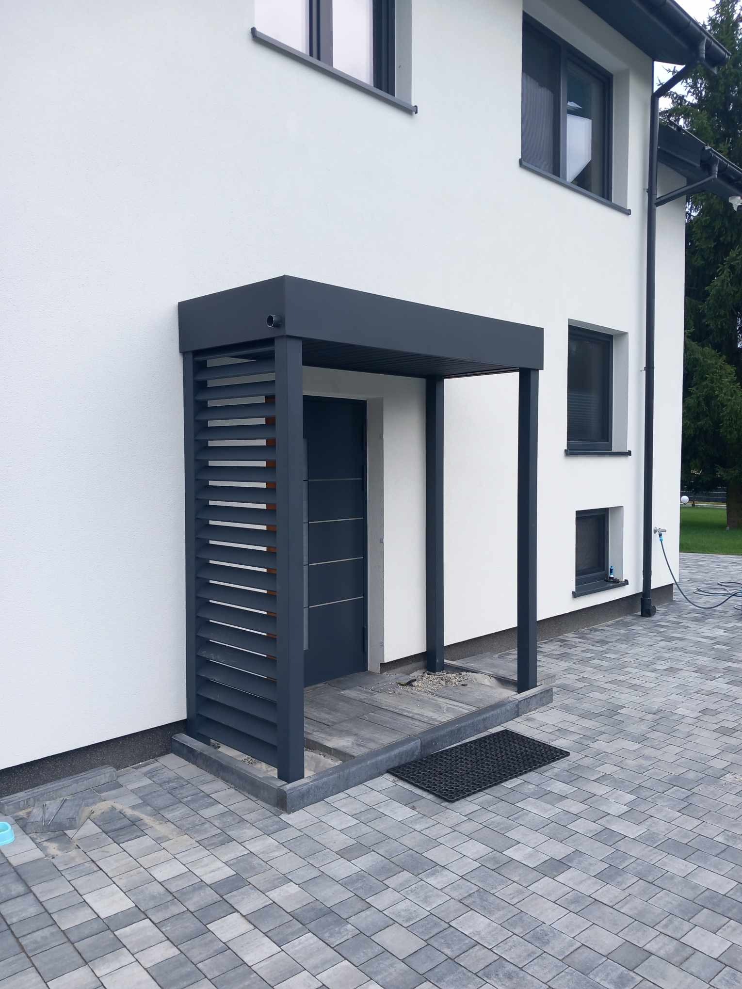 Nowoczesne zadaszenie nad wejściem do domu, z metalową konstrukcją w kolorze grafitowym i lamelami po boku, dopasowane do minimalistycznej architektury budynku.