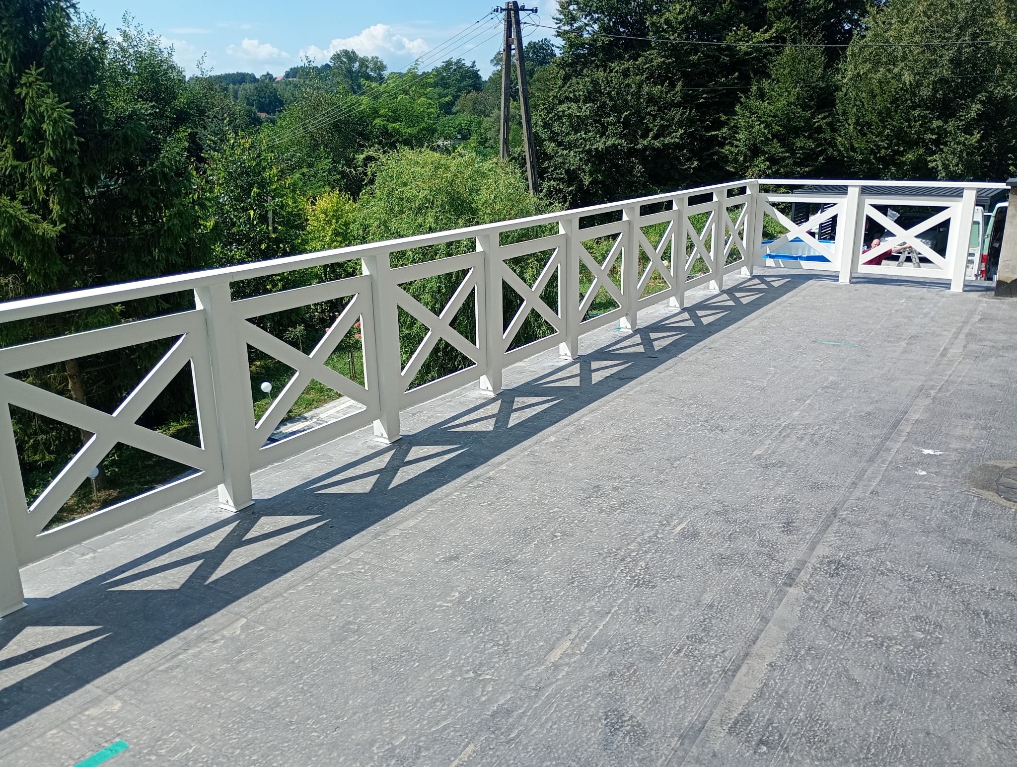 Biała balustrada z krzyżowym wzorem na tarasie z widokiem na zieleń, wyraźne cienie w słoneczny dzień.