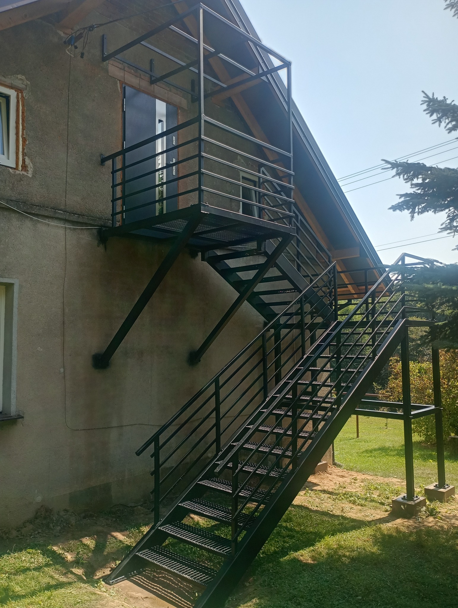 Zewnętrzne schody metalowe z balustradą prowadzące do drzwi na piętrze budynku z szarą elewacją.