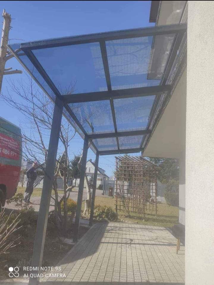 Metalowa konstrukcja pergoli z przezroczystym zadaszeniem, widoczna z poziomu chodnika, z fragmentem czerwonego samochodu dostawczego w tle.