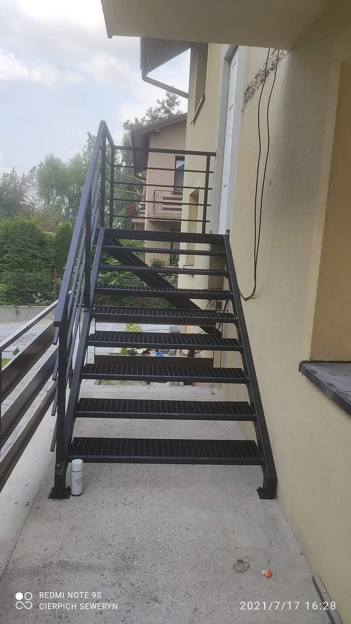 Zewnętrzne schody metalowe z czarnego profilu, prowadzące na balkon, z widoczną puszką farby w tle.