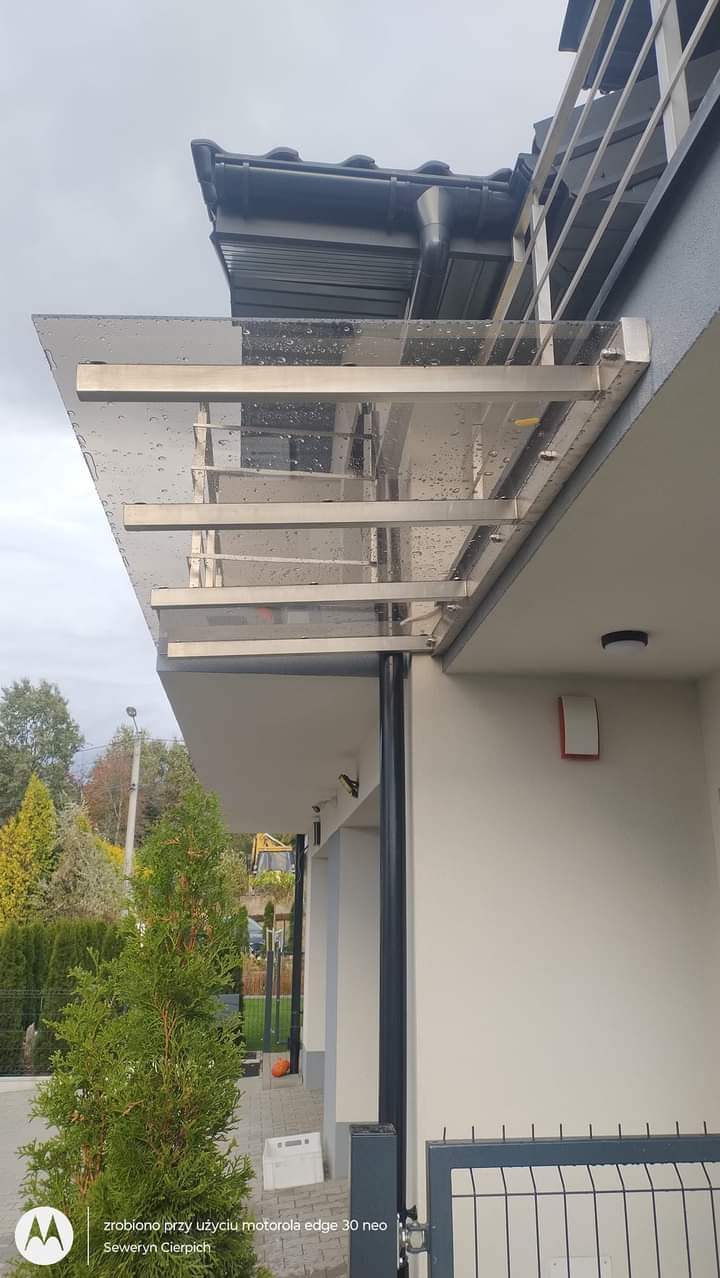 Zadaszenie nad wejściem do domu, konstrukcja ze stali nierdzewnej i przezroczystego tworzywa, widoczne krople deszczu na powierzchni, w tle drzewa i fragment ogrodzenia panelowego.