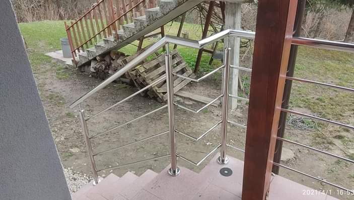 Zewnętrzna klatka schodowa z balustradą ze stali nierdzewnej i drewnianym słupkiem, widok z góry na schody i fragment podwórka z drewnem ułożonym obok starych schodów.