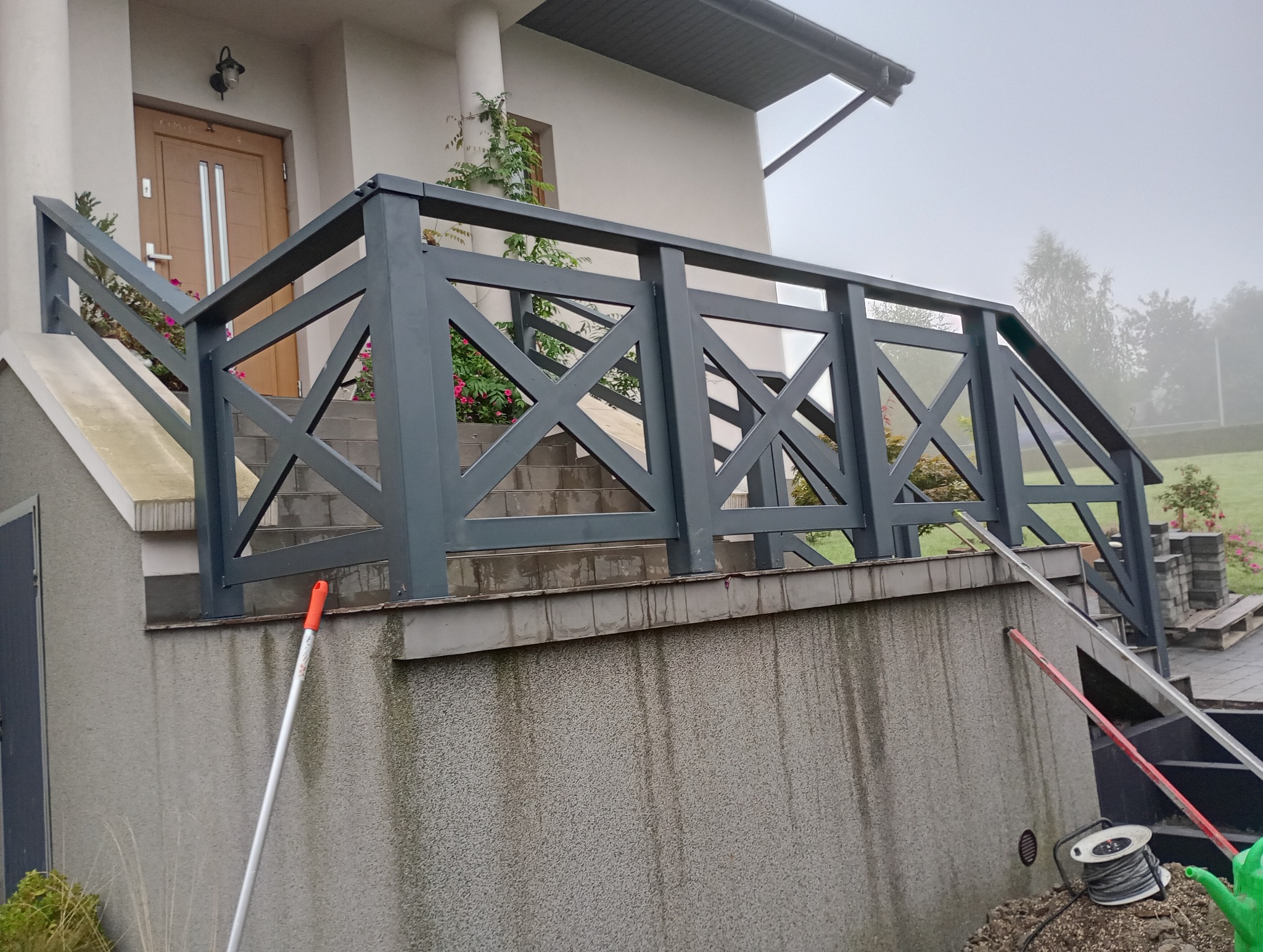 Grafitowa balustrada schodowa z krzyżowym wzorem na betonowym podeście, widoczne narzędzia i drabina sugerują prace montażowe.