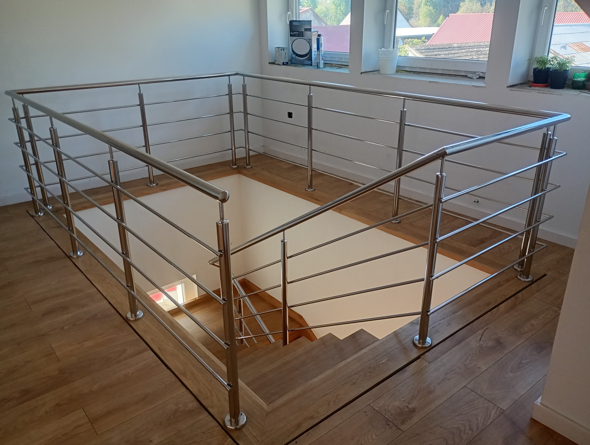 Nowoczesna balustrada ze stali nierdzewnej z poziomymi prętami, zabezpieczająca schody i antresolę, widok z góry na drewnianą podłogę i okno z roślinami.