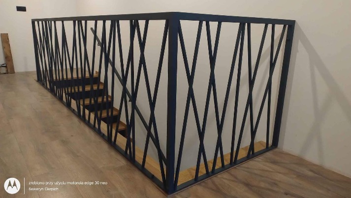 Nowoczesna, czarna balustrada metalowa z ukośnymi prętami zabezpieczająca schody drewniane wewnątrz domu.