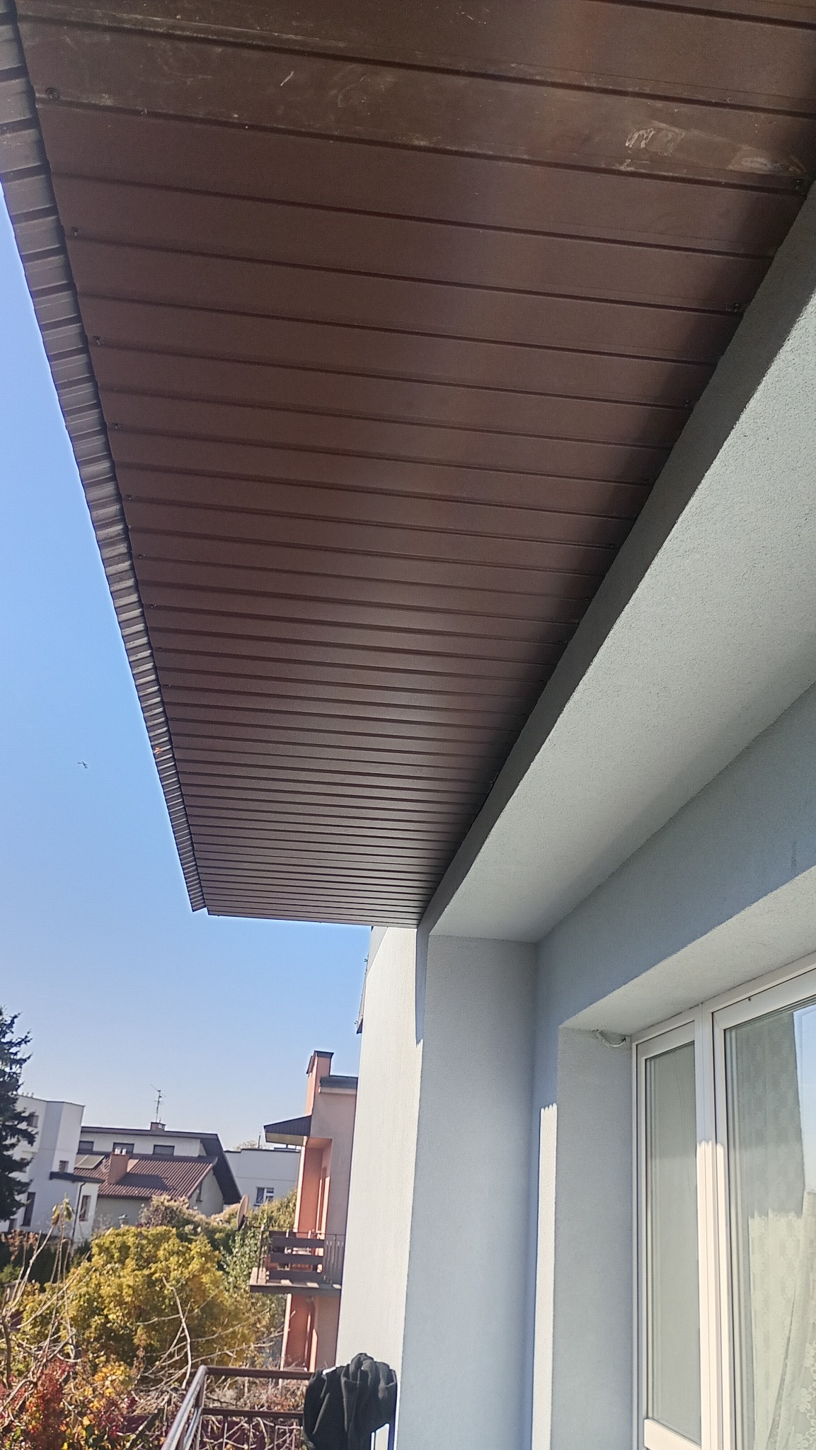 Podbitka dachowa z brązowych paneli PCV zamontowana na balkonie budynku mieszkalnego, widok z dołu na tle błękitnego nieba i zabudowy miejskiej.