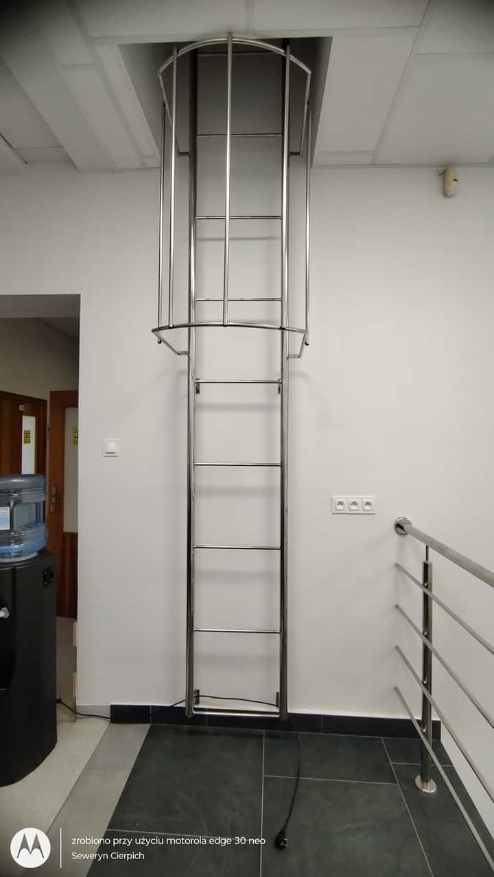 Zamontowana w Tarnowie drabina ewakuacyjna ze stali nierdzewnej, prowadząca do otworu w suficie, z zabezpieczającym koszem. Widoczne fragmenty balustrady i wyposażenia biurowego.
