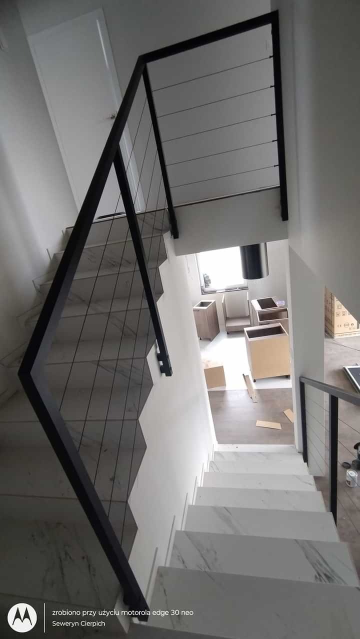 Wewnętrzne schody wykończone białym marmurem z czarną, minimalistyczną balustradą z cienkich, poziomych linek, widziane z góry w trakcie montażu zabudowy kuchennej.