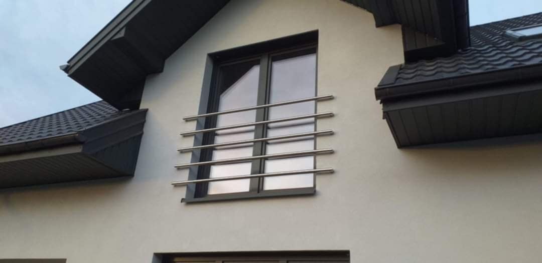 Zewnętrzna balustrada okienna z polerowanej stali nierdzewnej, składająca się z czterech poziomych prętów zamocowanych na elewacji budynku z ciemną stolarką okienną i grafitowym dachem.