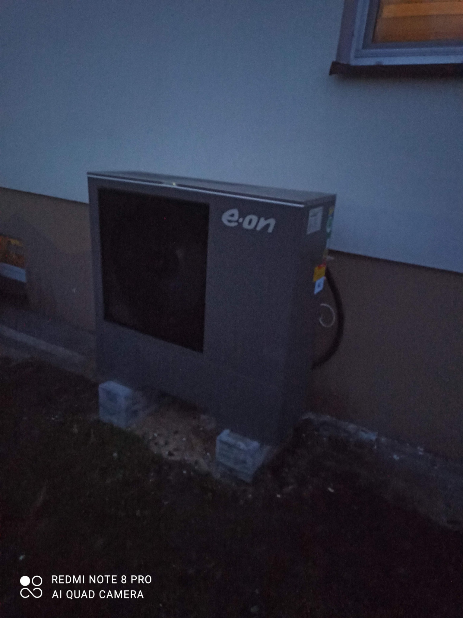 Szara pompa ciepła E-on, zamontowana na betonowych bloczkach przy ścianie budynku. Widoczne przewody i fragment okna. Zdjęcie wykonane o zmierzchu.