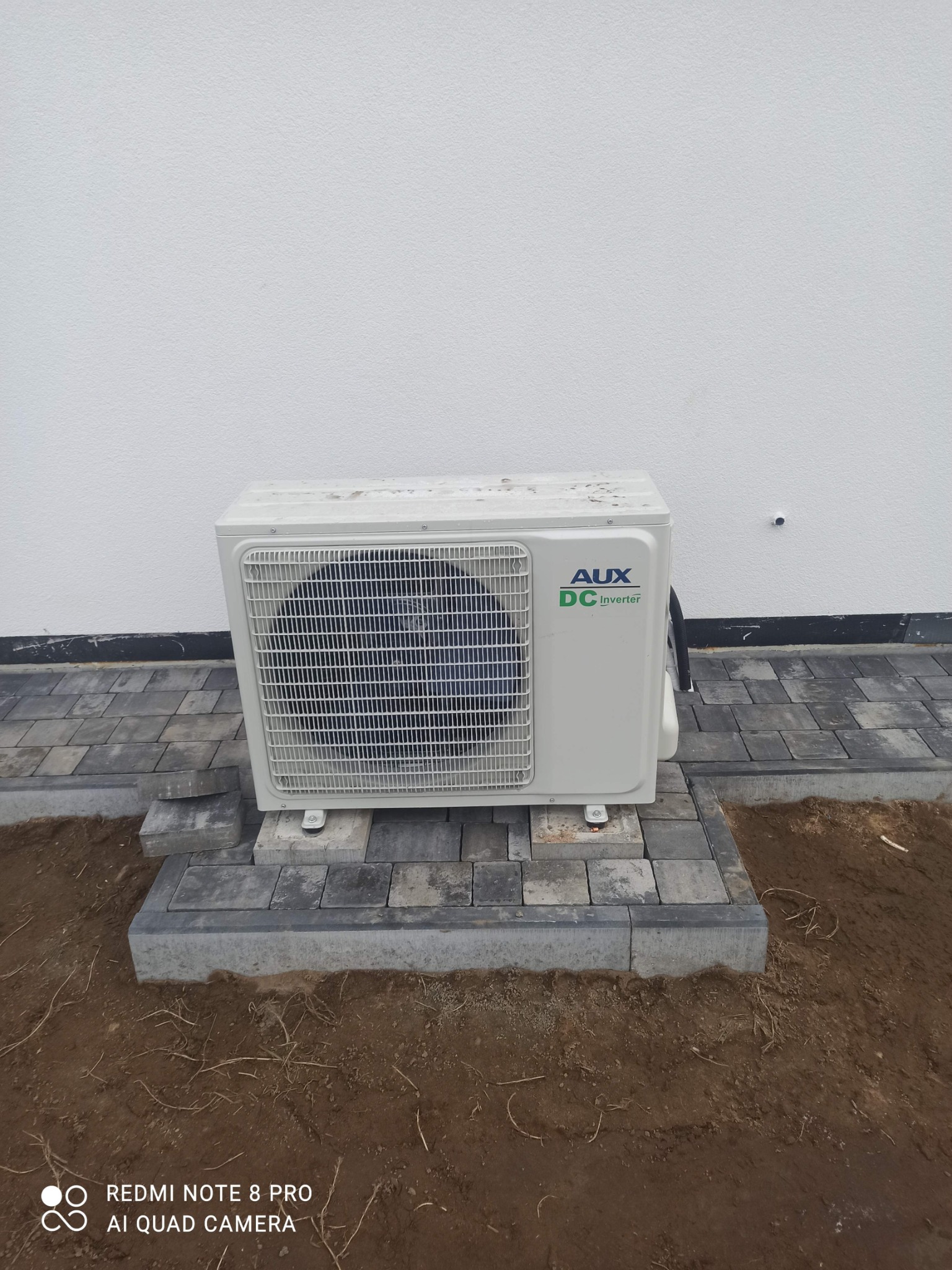 Biała zewnętrzna jednostka klimatyzacji AUX DC Inverter, zamontowana na szarych betonowych płytach chodnikowych przy białej ścianie budynku.