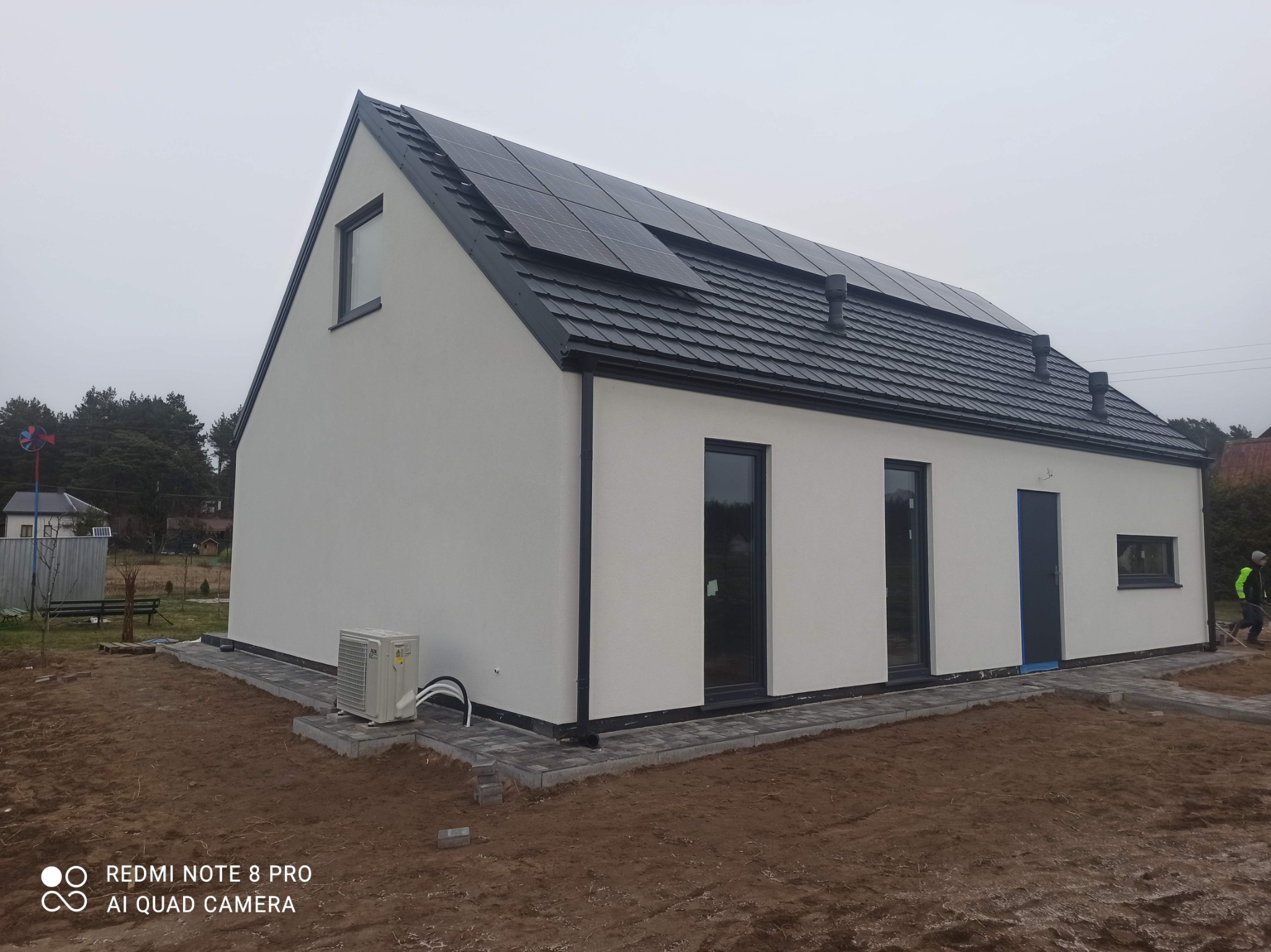 Biały dom jednorodzinny z panelami słonecznymi na dachu i klimatyzatorem na zewnątrz. Widoczne okna z ciemnymi ramami i kostka brukowa wokół budynku.