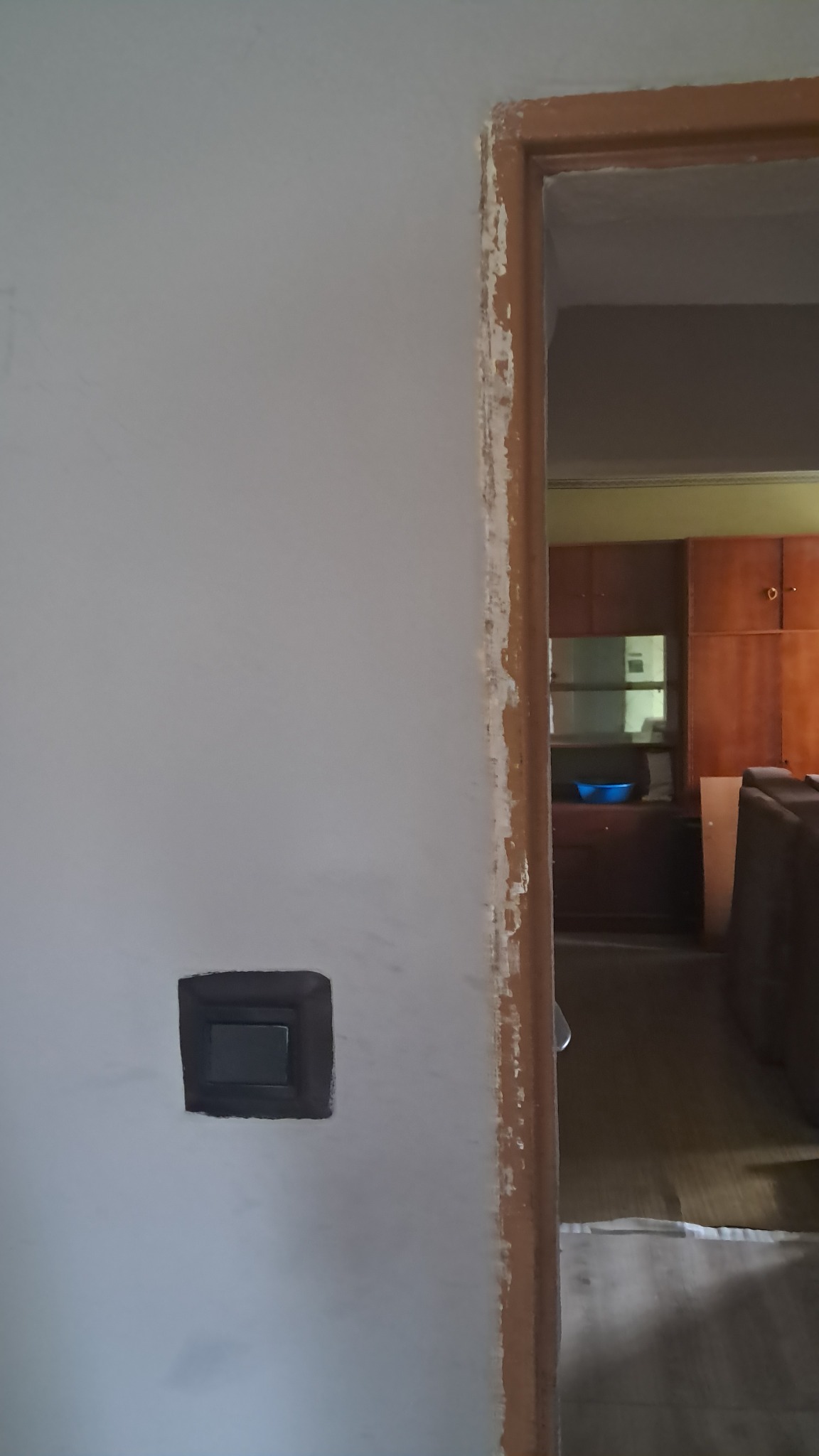 Przygotowanie ściany do malowania: Ościeżnica drzwi z resztkami starej farby i szpachli, obok szary włącznik światła na gładkiej, jasnoszarej ścianie.