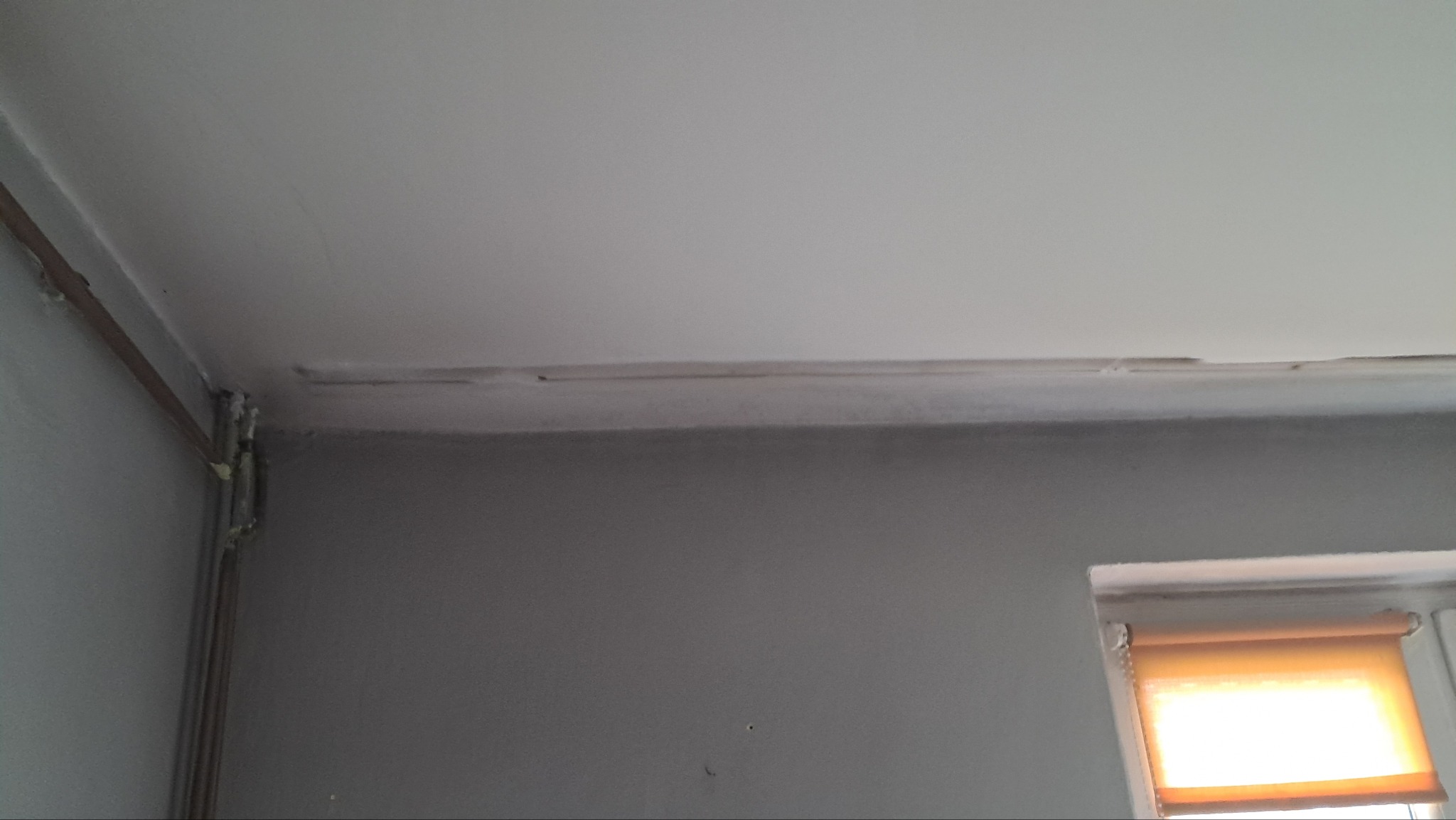 Szare wnętrze pokoju z widocznym fragmentem sufitu po naprawie, gdzie widać ślady szpachli i nierówności, a także rury przy ścianie zabezpieczone pianką montażową, oraz fragment okna z roletą.