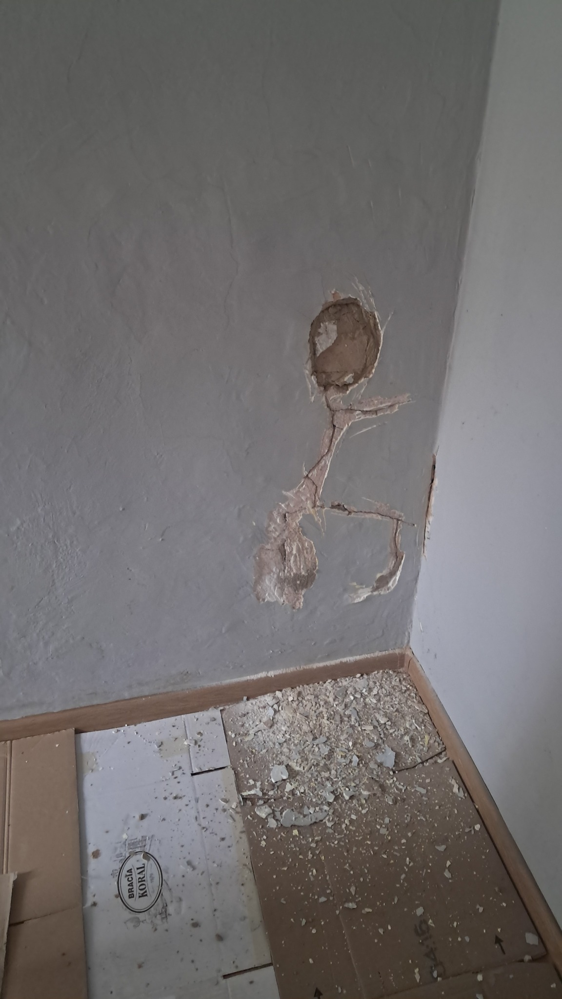 Uszkodzona ściana w rogu pokoju, z widocznymi ubytkami tynku i pęknięciami, zabezpieczona kartonem na podłodze, z rozsypanymi odłamkami tynku.