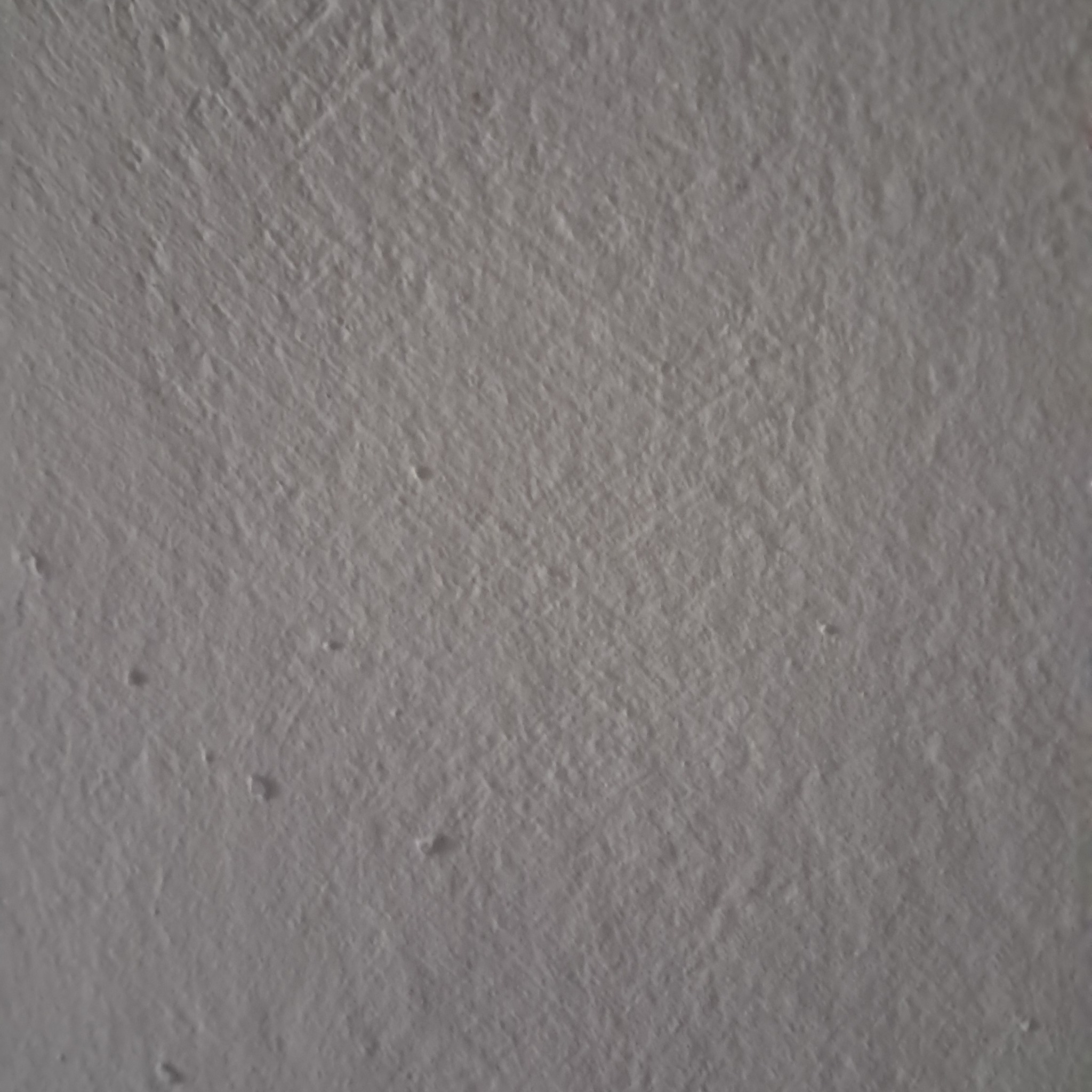 Szara, nierówna powierzchnia ściany z widocznymi drobnymi nierównościami i rysami, oświetlona z góry.