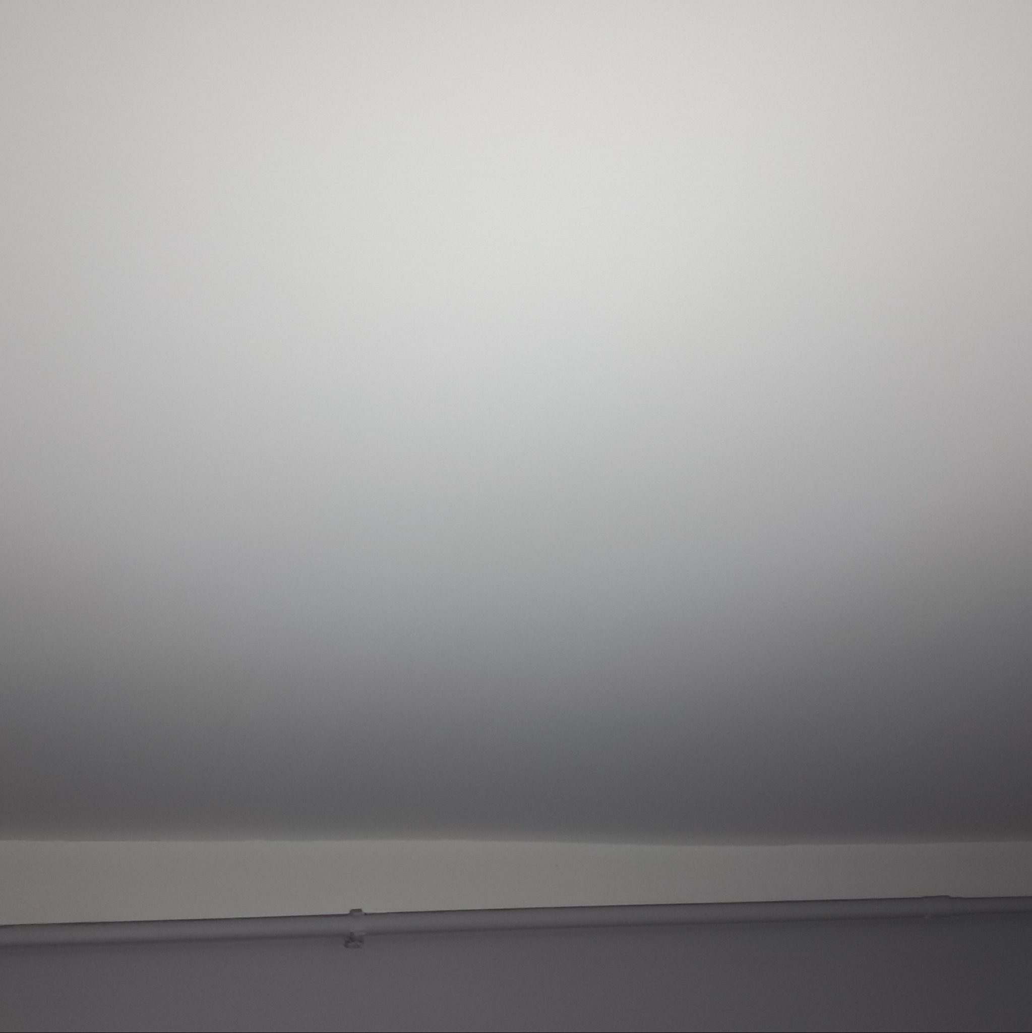 Fragment sufitu z widoczną, pomalowaną na biało listwą przypodłogową i fragmentem rury instalacyjnej. Sufit ma nierównomierne oświetlenie, przechodzące od jaśniejszego u góry do ciemniejszego...