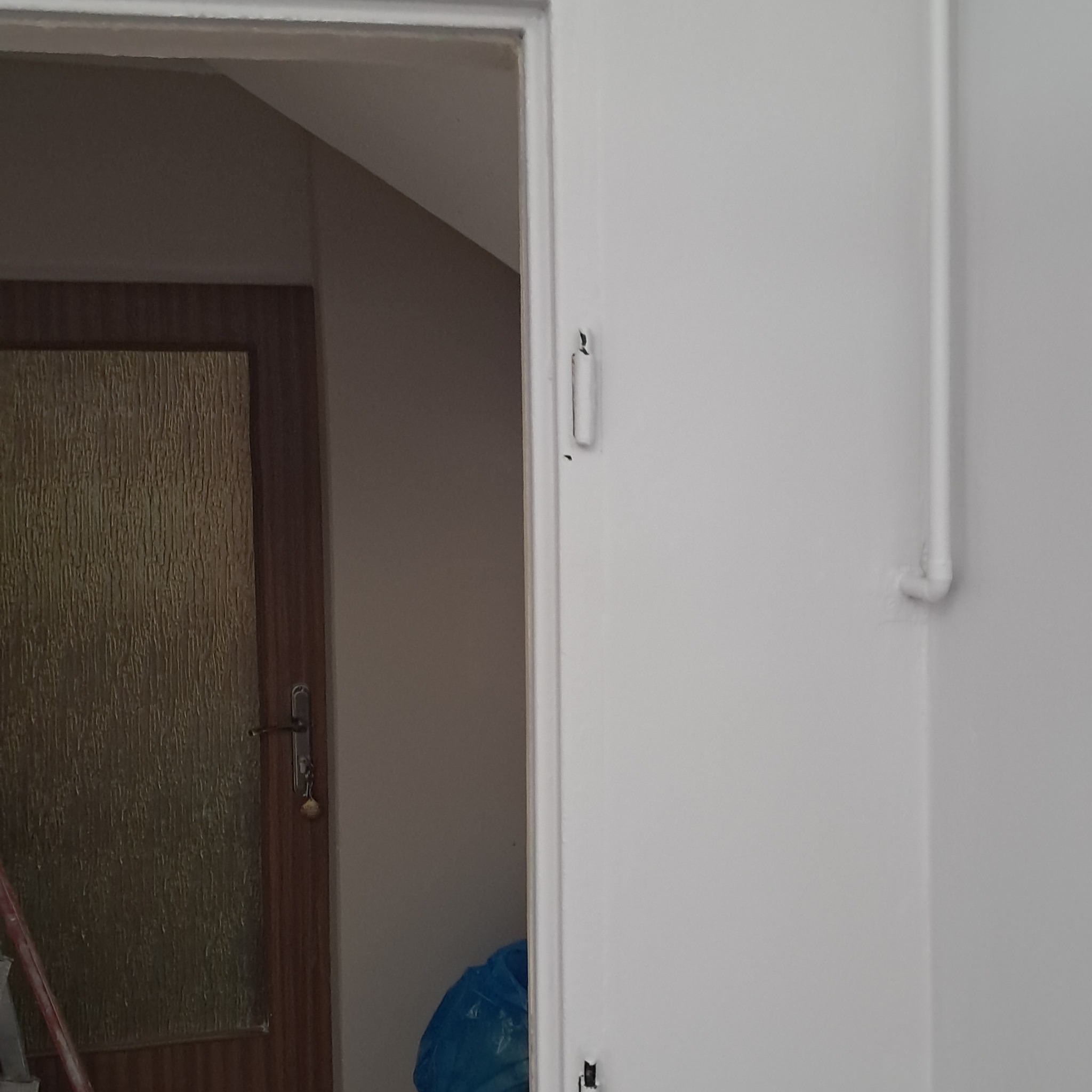 Biała ściana po świeżym malowaniu z widoczną rurą instalacyjną i fragmentem drzwi z szybą w tle.