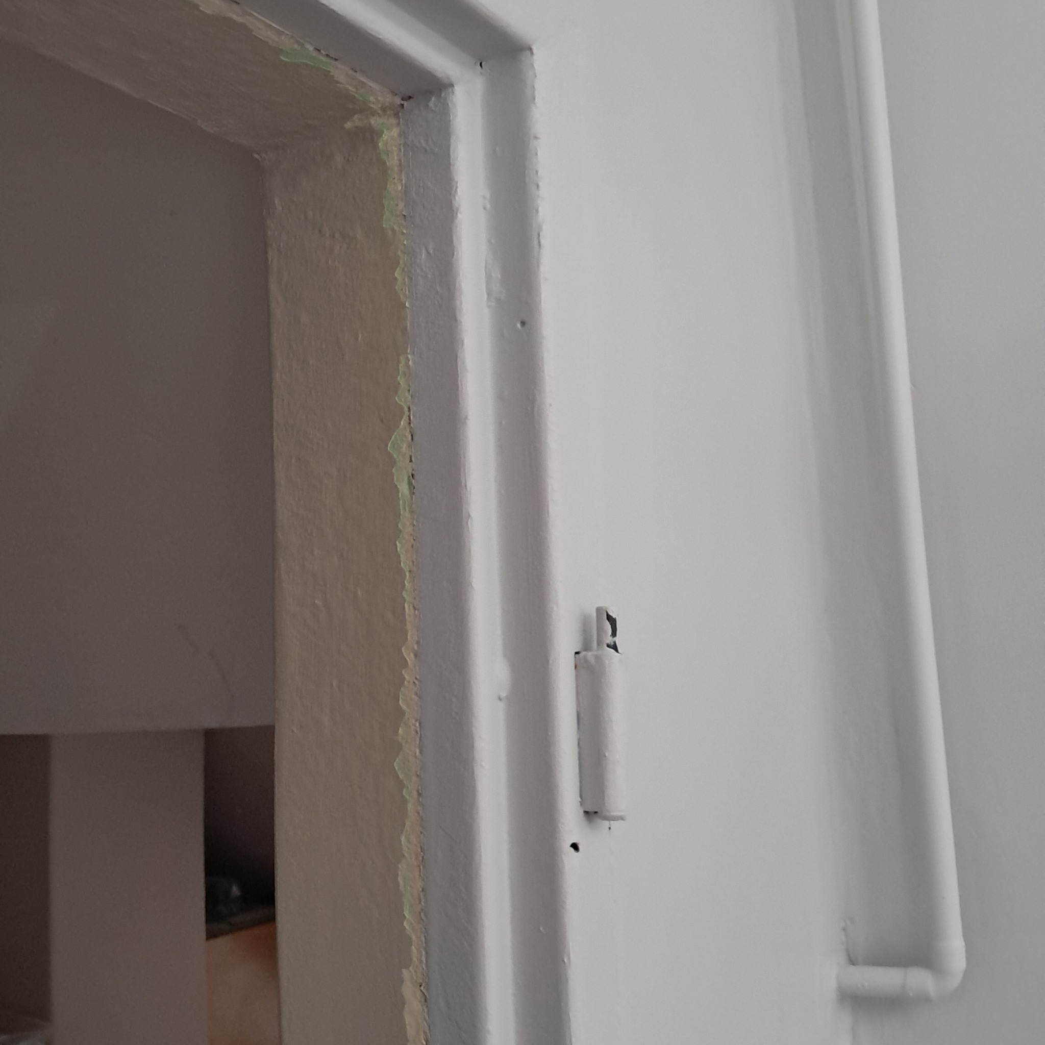 Fragment ościeżnicy drzwiowej w trakcie prac wykończeniowych, widoczne ślady starej farby i nierówności na powierzchni przygotowywanej do malowania, obok pionowy element instalacji pomalowany...