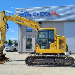 CHOGI SPÓŁKA Z OGRANICZONĄ ODPOWIEDZIALNOŚCIĄ - Żółta koparka Komatsu z łyżką na tle budynku z logo firmy, widok z boku, na pierwszym planie kostka brukowa, w tle zaparkowane samochody.
